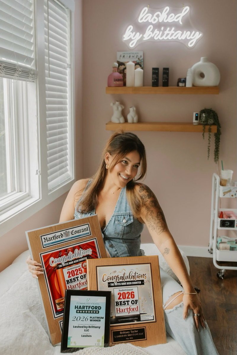 Brittany seated in her studio holding framed Best of Hartford award plaques, with the neon "lashed by brittany" sign lit behind her
