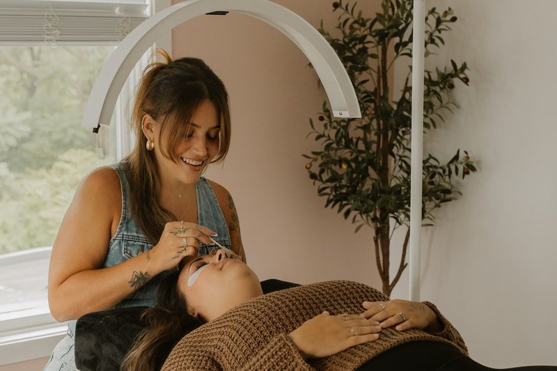 The treatment room: a client reclining under a ring light beside a bright window and a potted tree