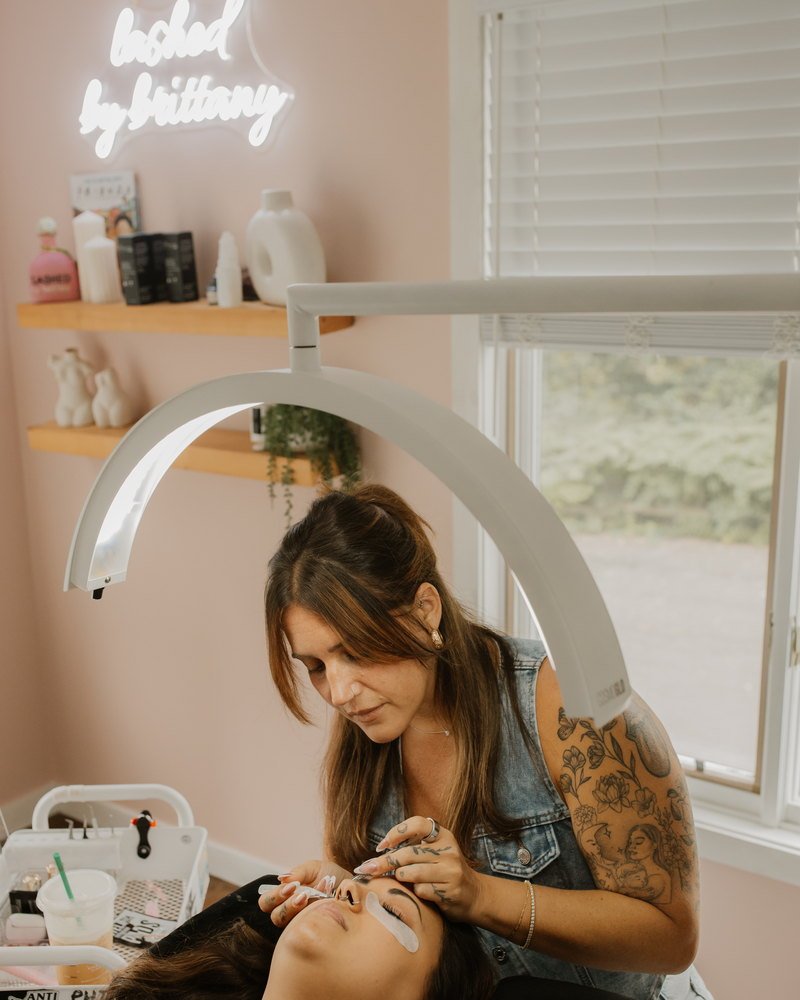 Brittany applying lash extensions beneath a ring light, with the studio's neon "lashed by brittany" sign on the wall behind her